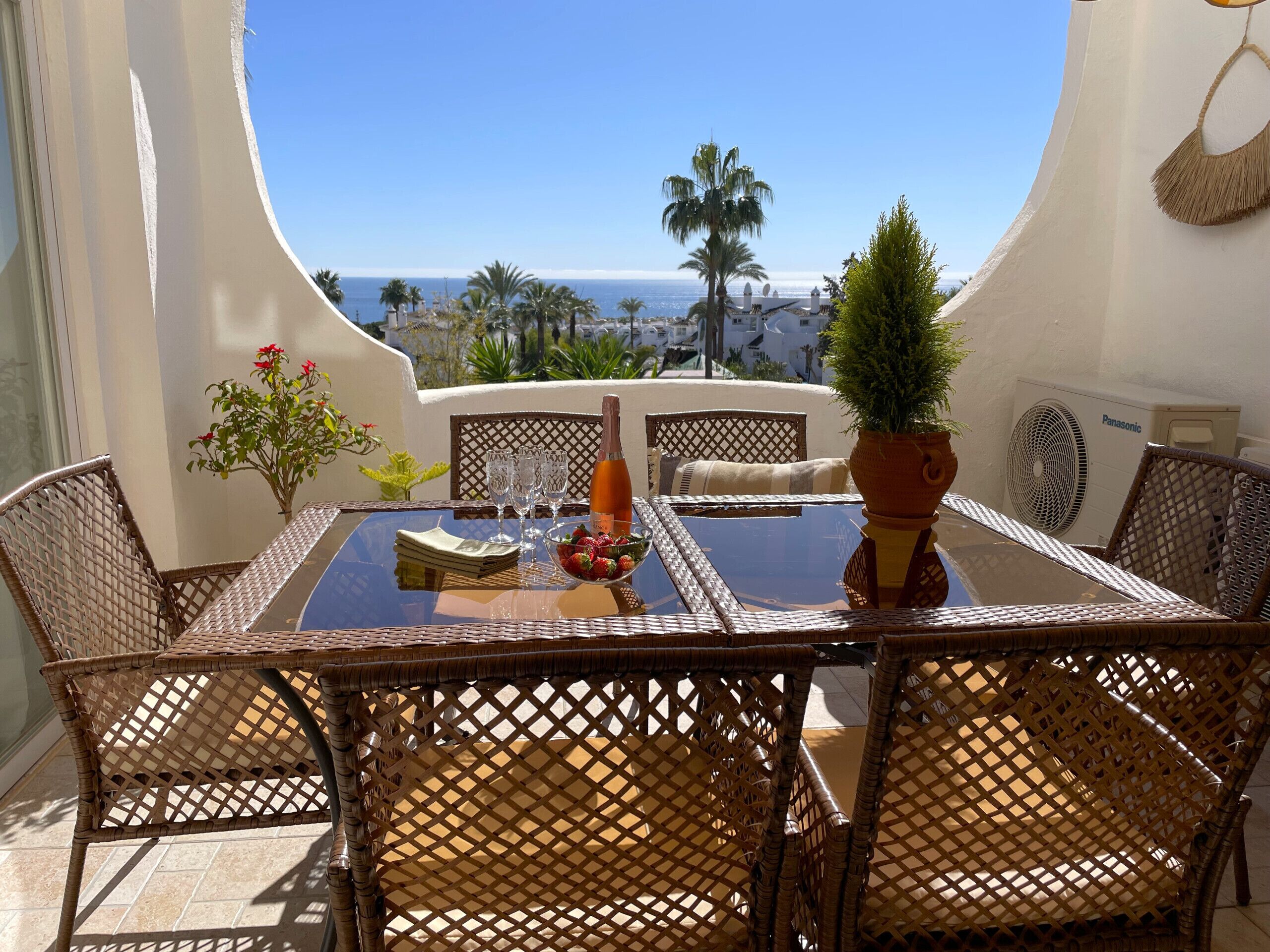 A dining area with wicker chairs and a glass table, overlooking a coastal view with palm trees and a clear sky.