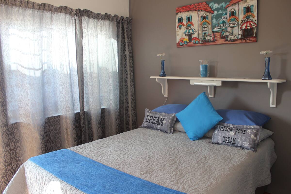 Una cama bien hecha con un edredón azul, dos almohadas con el texto 'DREAM' y una almohada azul, además de un estante de pared con jarrones.