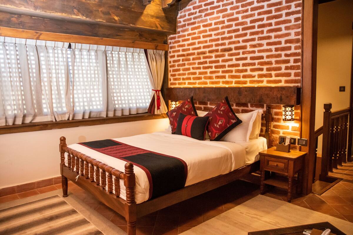 A wooden bed with a brick wall behind it, a window with curtains, and a nightstand with a lamp.