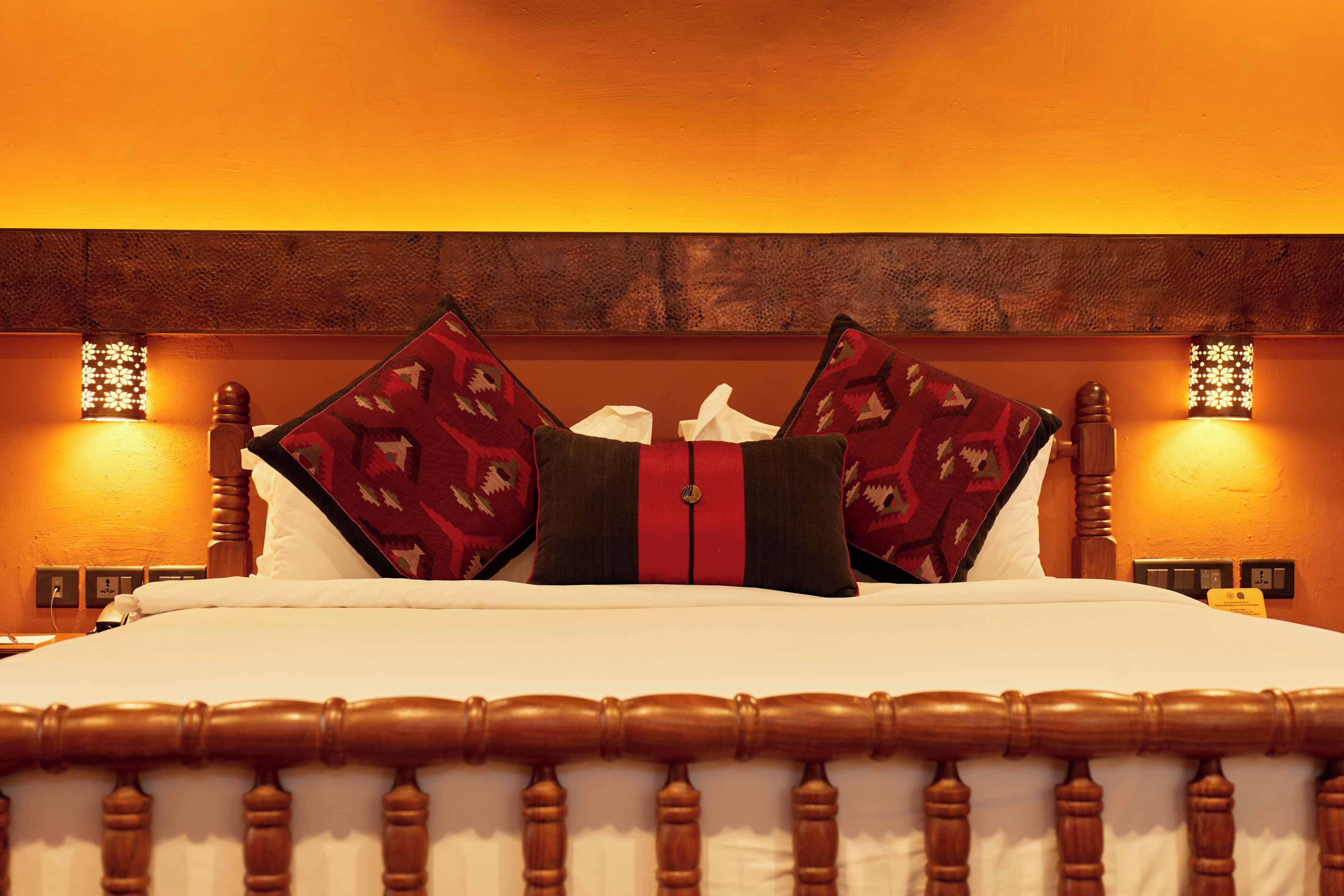 Una cama con cabecera de madera, dos lámparas fijadas a la pared y almohadas estampadas.