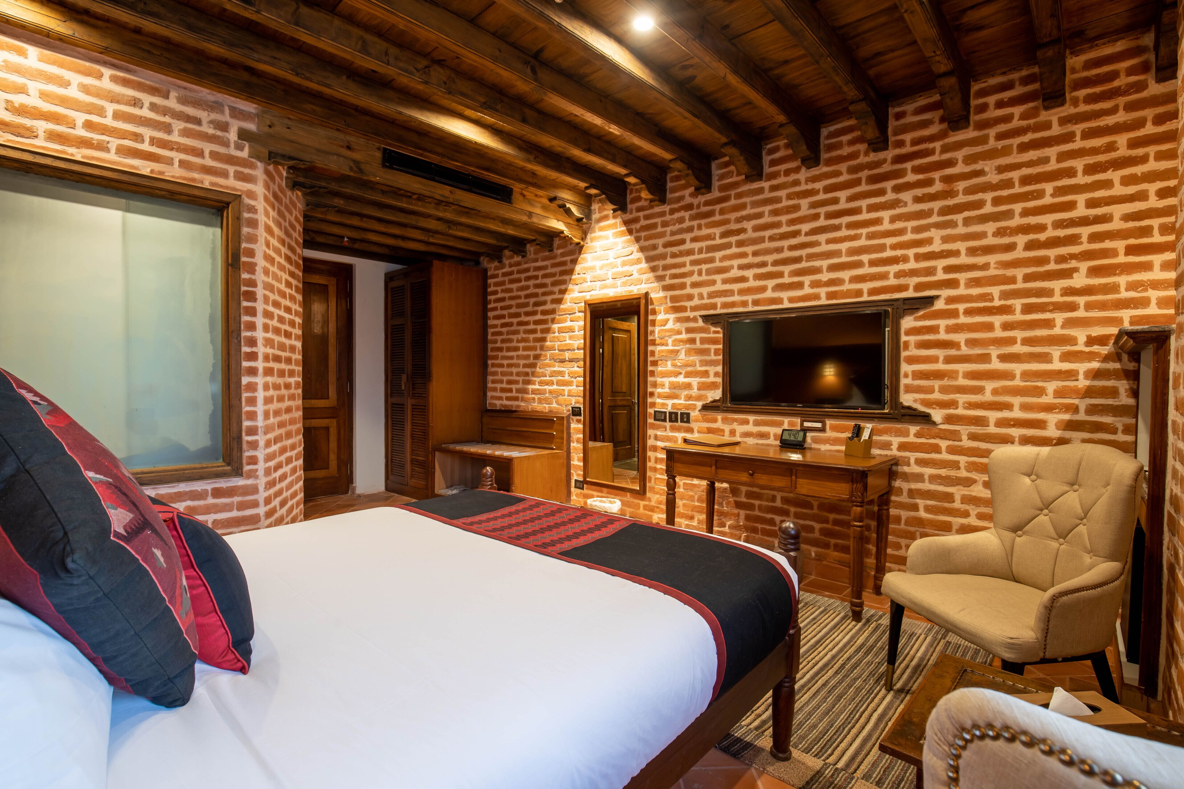 Un dormitorio con una cama grande, techo de madera, pared de ladrillo, televisor, una silla y un escritorio.
