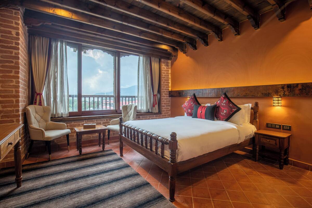 A bedroom with a large bed, a wooden headboard, a nightstand, a chair, a small table, and a view of the outdoors through a window.
