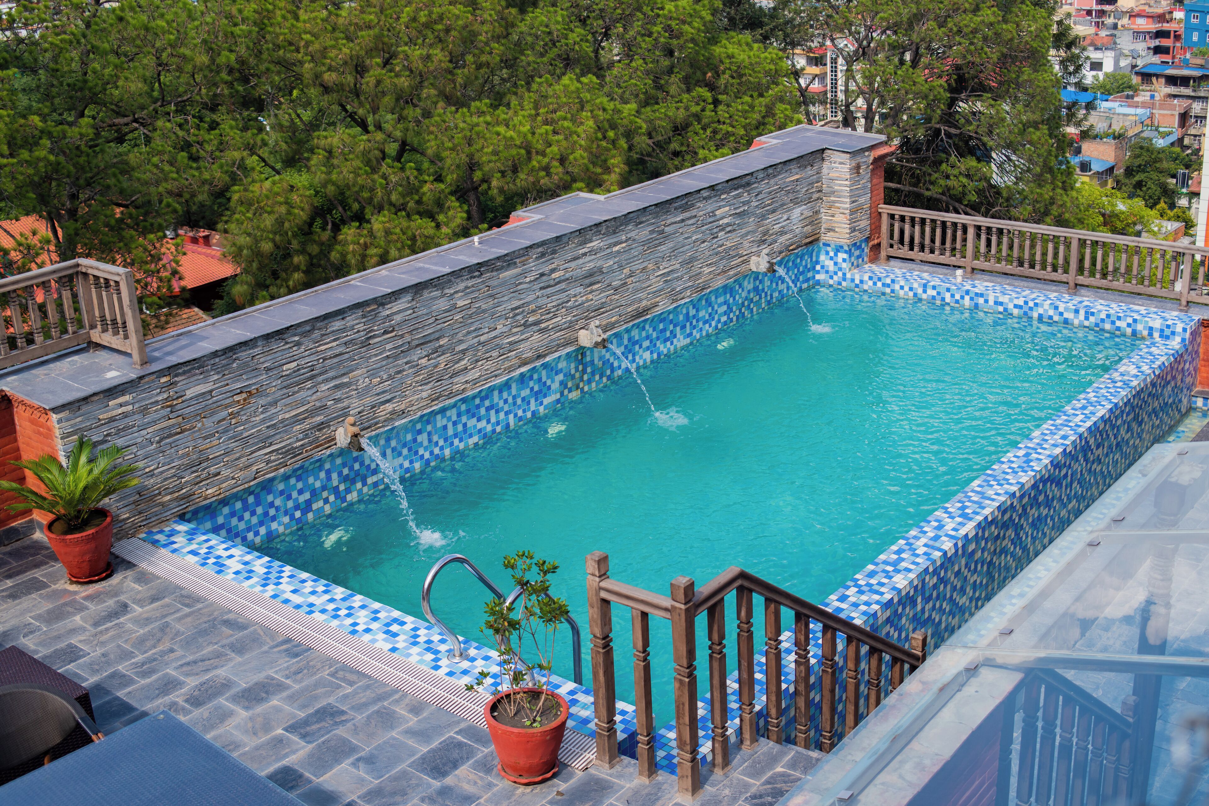Une piscine sur le toit, avec des murs en pierre et des bords recouverts de carreaux bleus, entourée de plantes en pot et d’une rambarde en bois.
