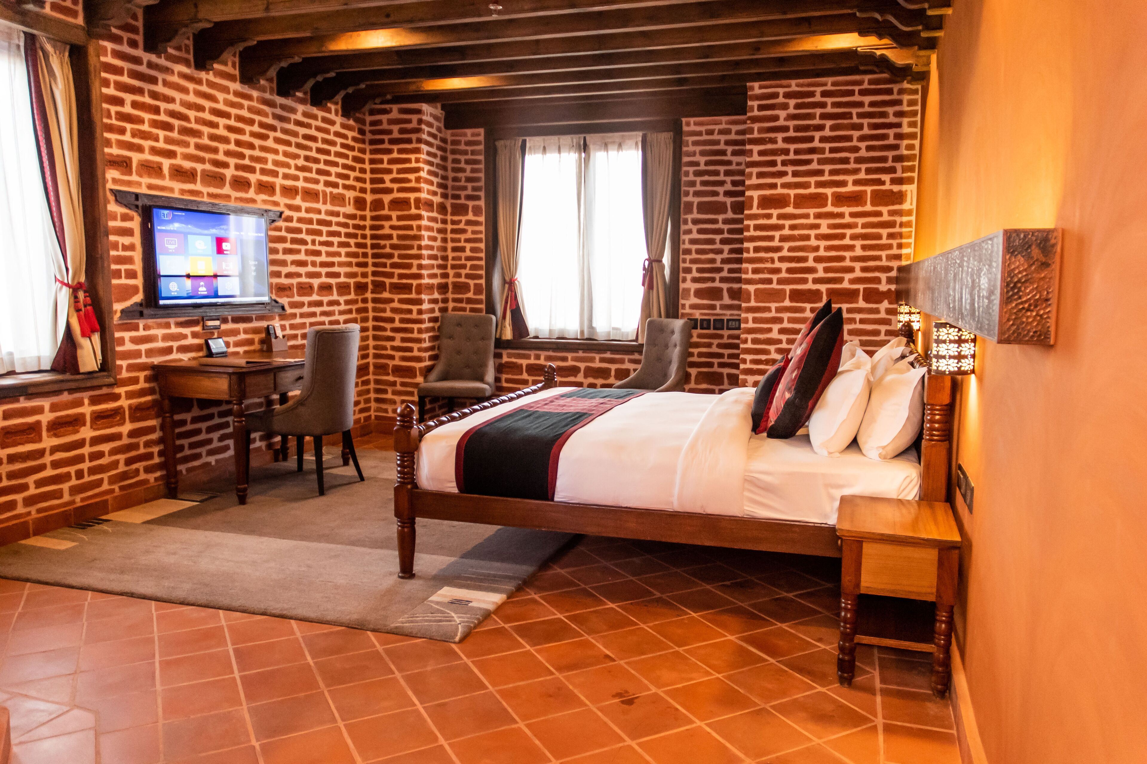 A hotel room with a brick wall, a bed, a desk with a computer, and a TV mounted on the wall.