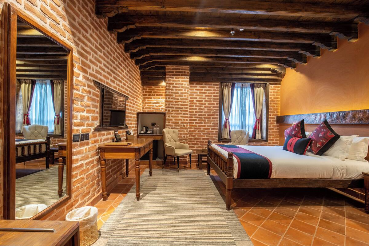 A room with a brick wall, wooden ceiling beams, a bed with a patterned bedspread, a desk with a chair, and a large mirror.