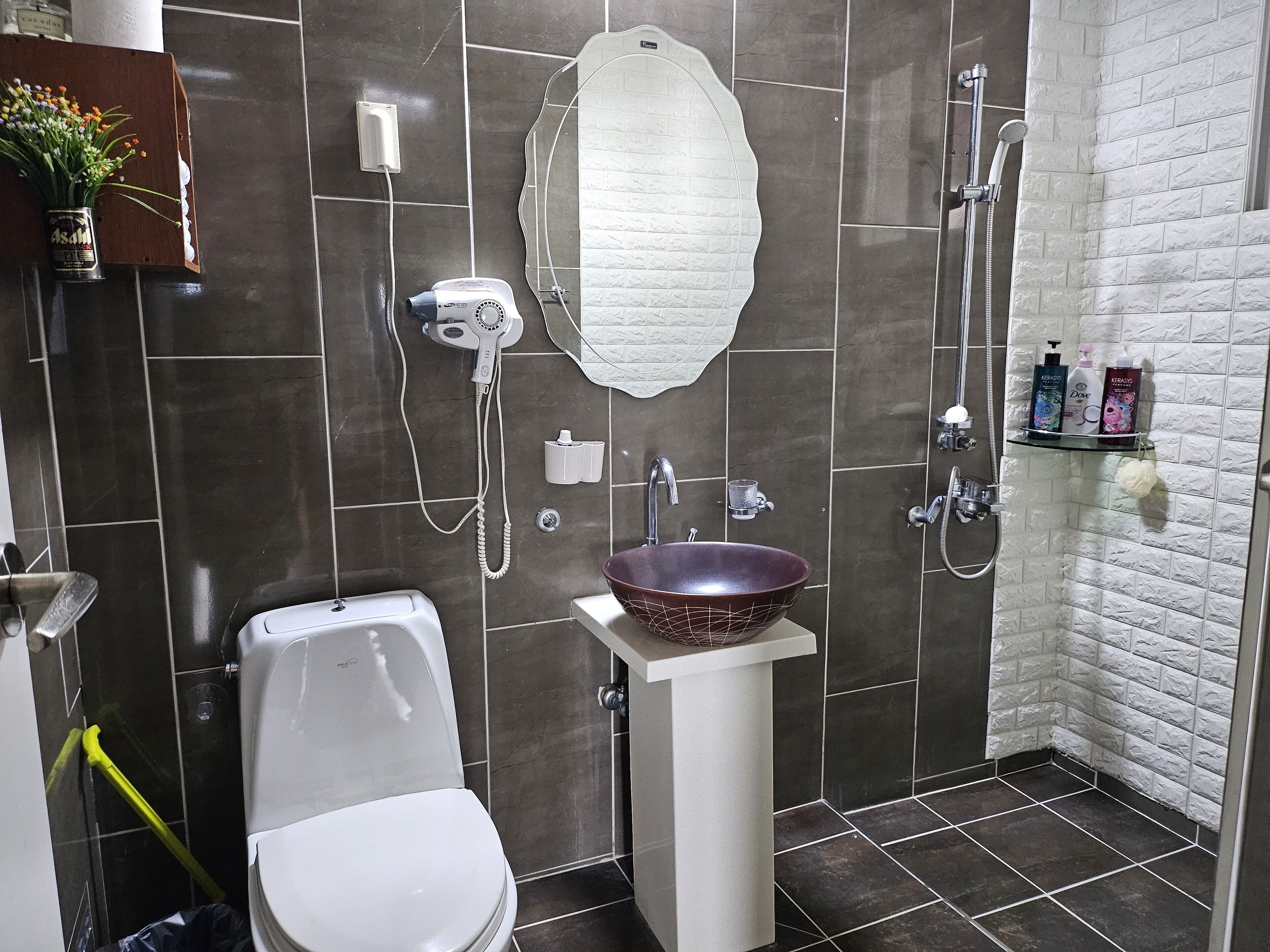 Un baño con inodoro blanco, un lavamanos blanco con un recipiente decorativo, un secador de pelo, una ducha con cabezal de ducha de mano y artículos de aseo en un estante.