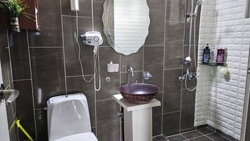 Un baño con inodoro blanco, un lavamanos blanco con un recipiente decorativo, un secador de pelo, una ducha con cabezal de ducha de mano y artículos de aseo en un estante.