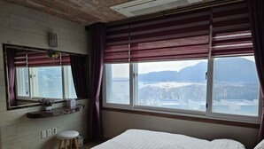 Une chambre avec un grand lit, une fenêtre donnant sur un plan d’eau et des collines, un petit tabouret et un miroir agrémenté d’une plante.