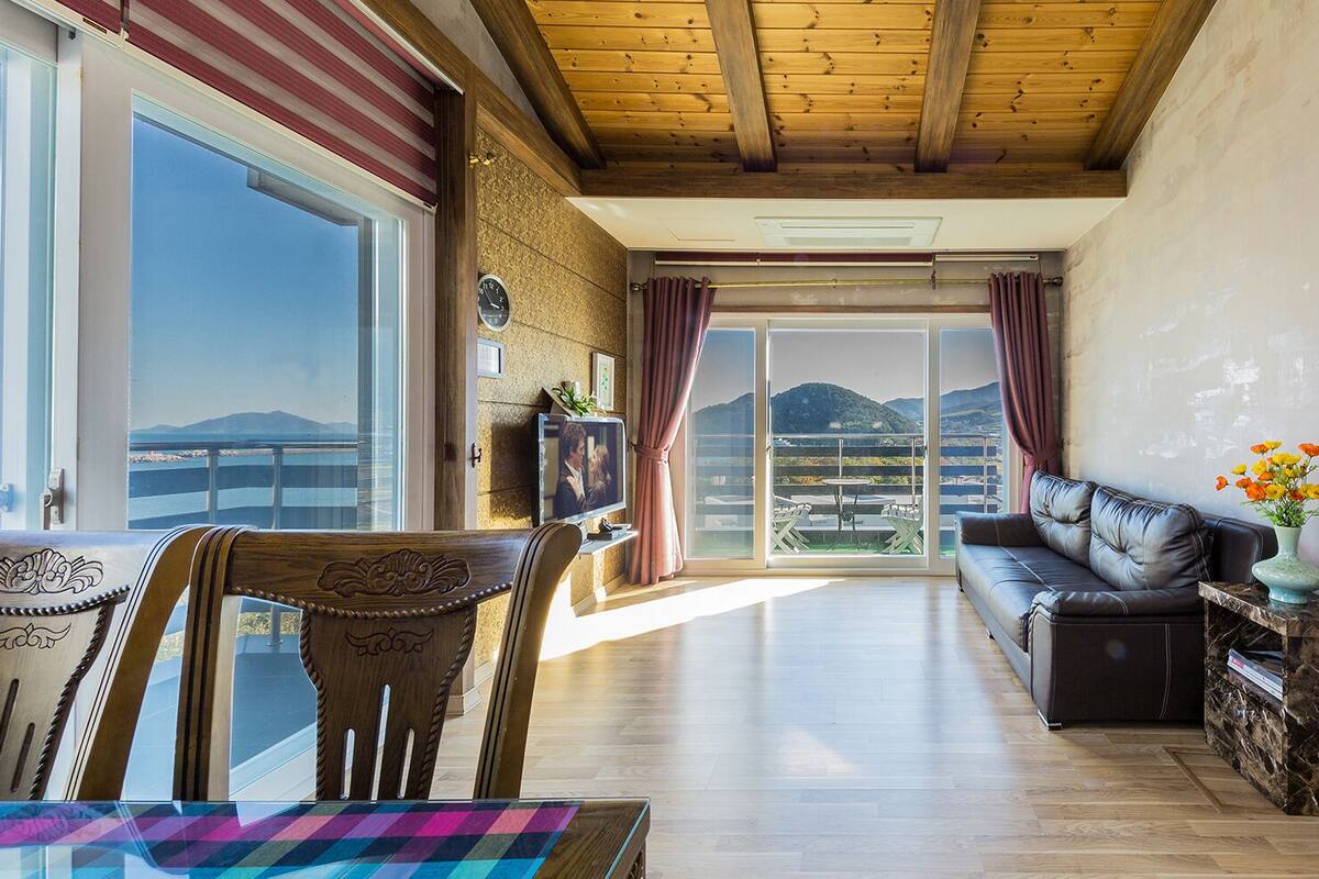 A spacious living room with a wooden ceiling, a large window, a sofa, a TV, and a view of mountains.