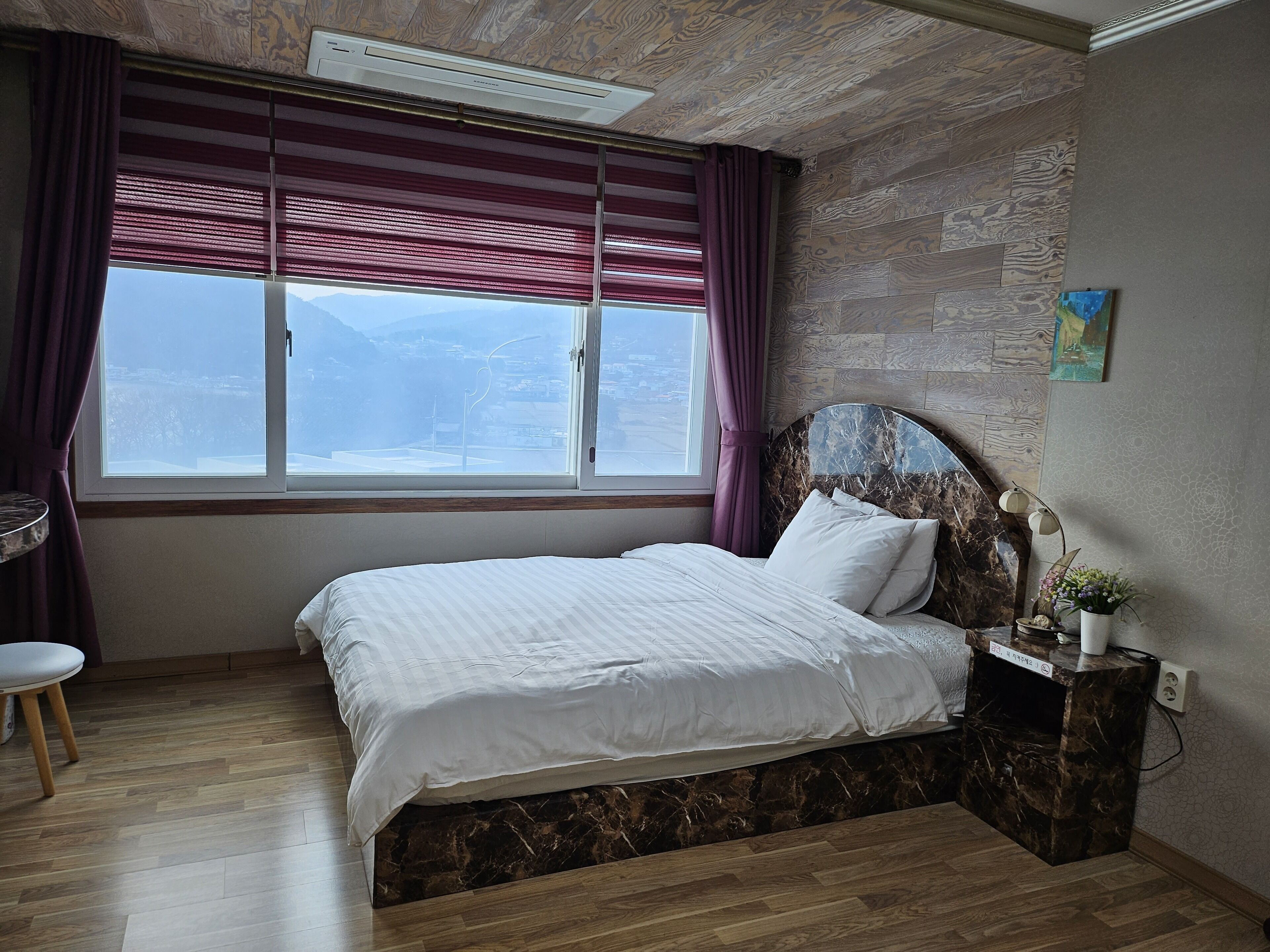 Een slaapkamer met een bed, gordijnen voor het raam, een hoofdeinde, een nachtkastje en een klein tafeltje met een stoel.