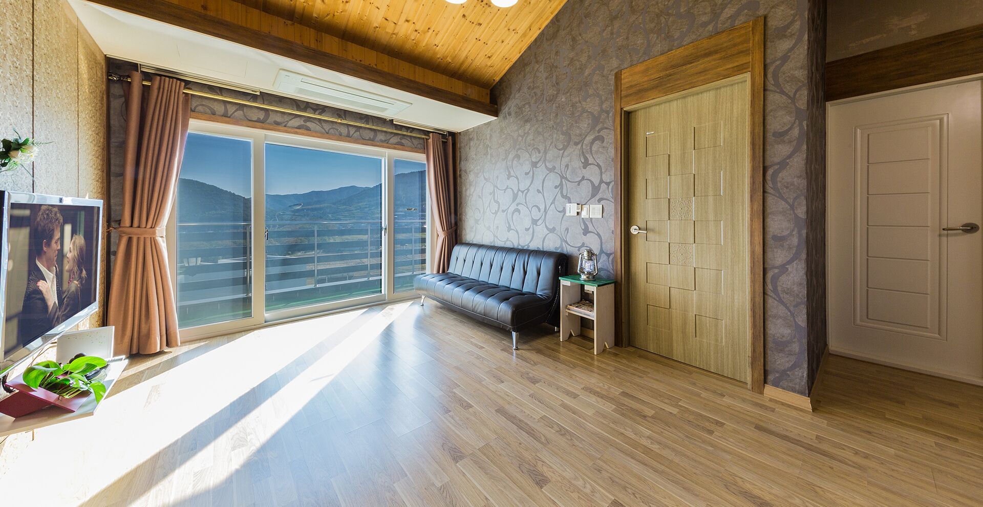 A spacious room with a large window, a sofa, a wooden door, and a television.