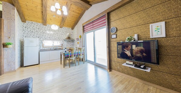 A modern kitchen with white cabinets, a dining area with a table and chairs, a television mounted on the wall, and a wooden ceiling. - Namhae Oceanview Pension (Namhae)