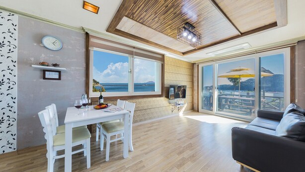 Un vaste espace de vie comprenant une table à manger, des chaises blanches, un canapé noir et de grandes fenêtres donnant sur la mer.