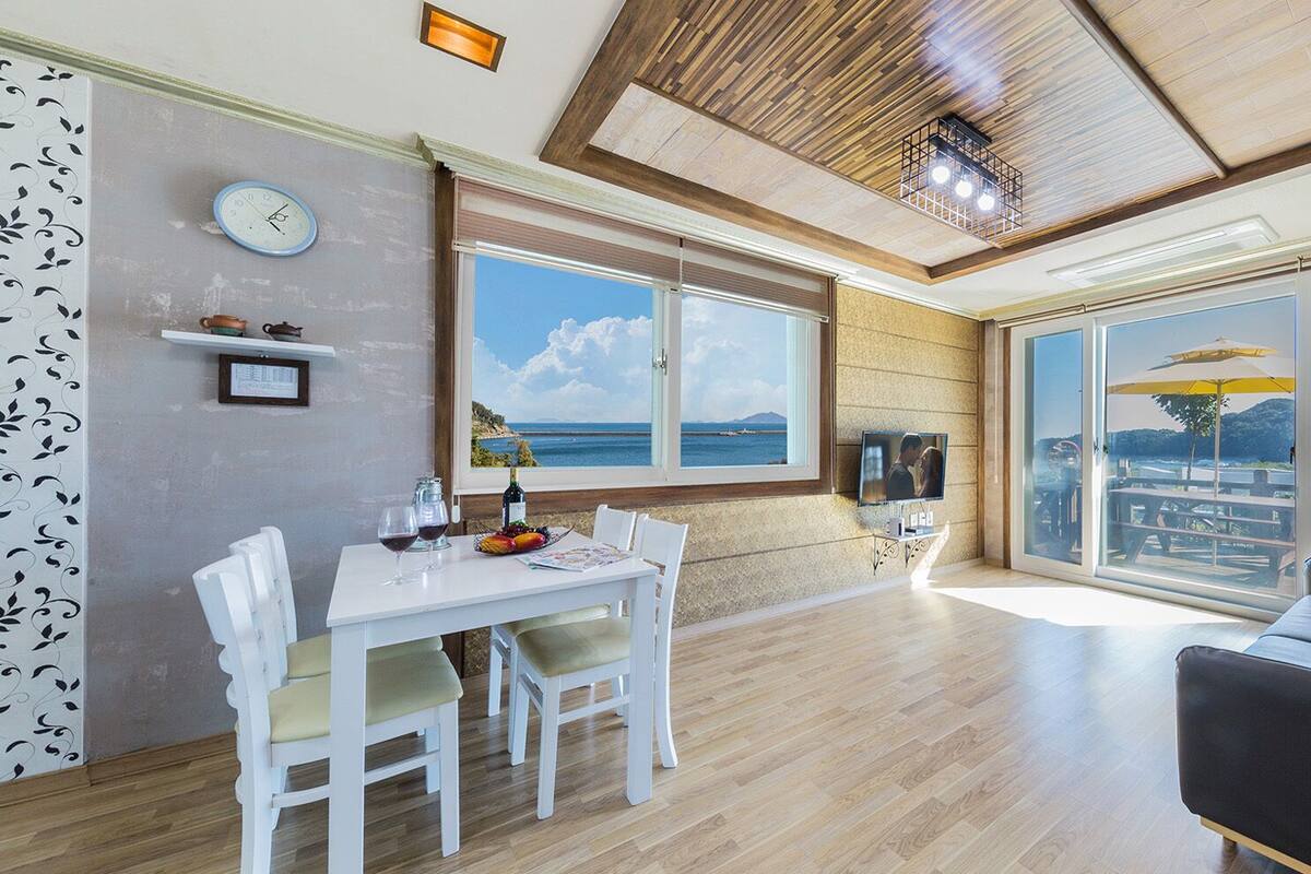 A spacious living area with a dining table, white chairs, a black sofa, and large windows offering a view of the sea.