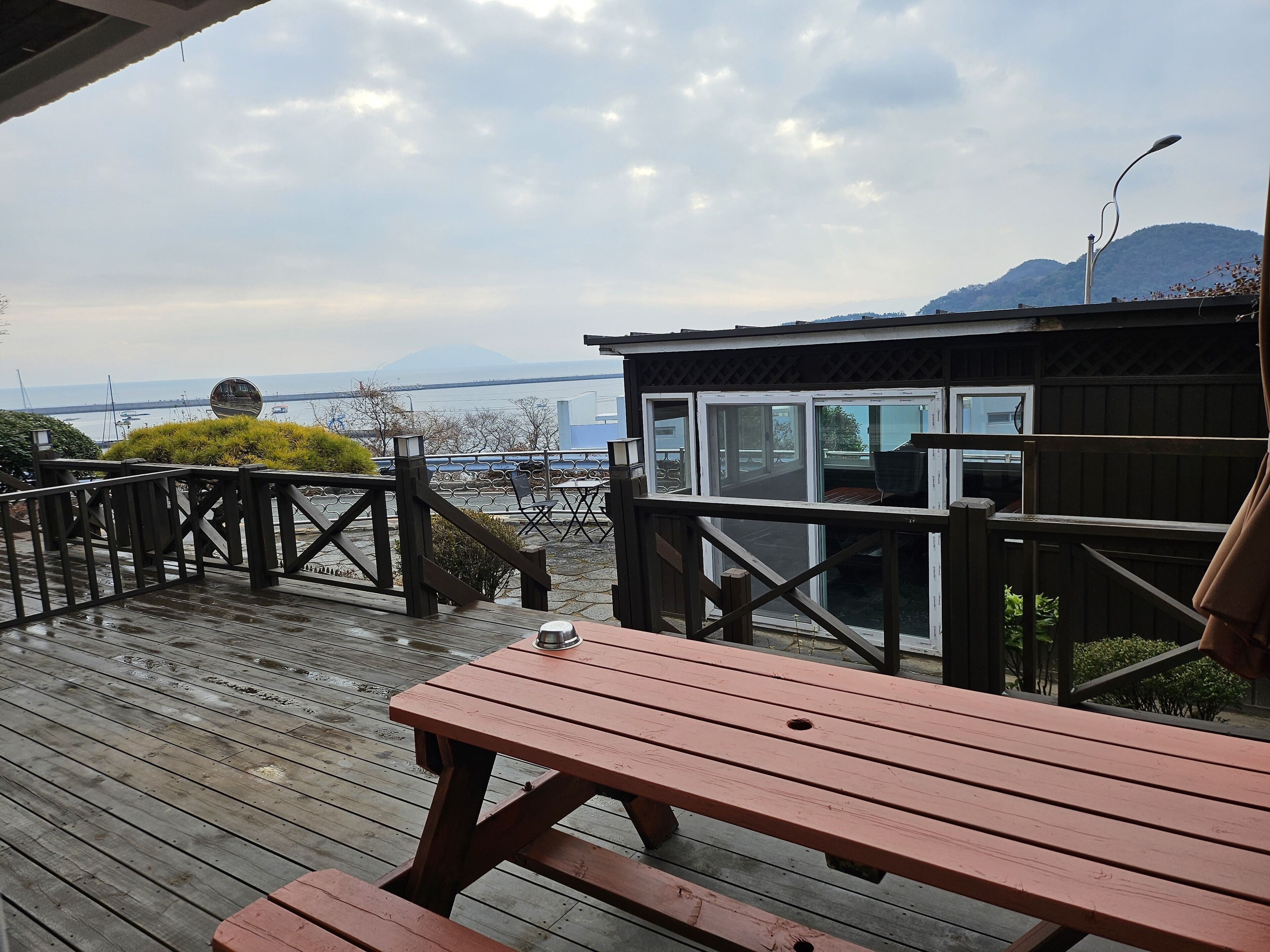A wooden deck with a picnic table and chairs, overlooking a body of water with boats and a distant mountain.
