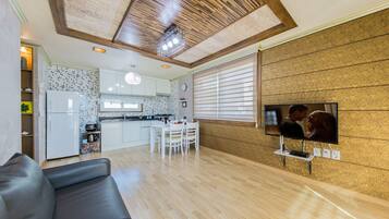 A modern kitchen with white cabinets, a dining area with a table and chairs, and a living room with a sofa and a TV mounted on the wall.