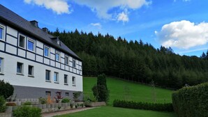 Un bâtiment à deux étages, doté d’une façade blanche et de moulures sombres, entouré d’une pelouse bien entretenue et bordé, à l’arrière, par une forêt dense.