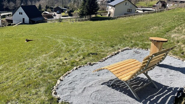 Un cadre rural avec un espace recouvert de gravier, comprenant un banc en bois et une petite table en bois, le tout entouré de maisons et d’une colline boisée.