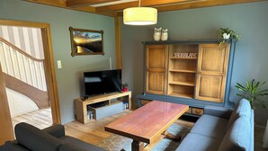 Un salon moderne avec un meuble TV en bois, un téléviseur à écran plat, un buffet en bois portant l’inscription « SAUERLAND », une table basse en bois et des canapés gris.