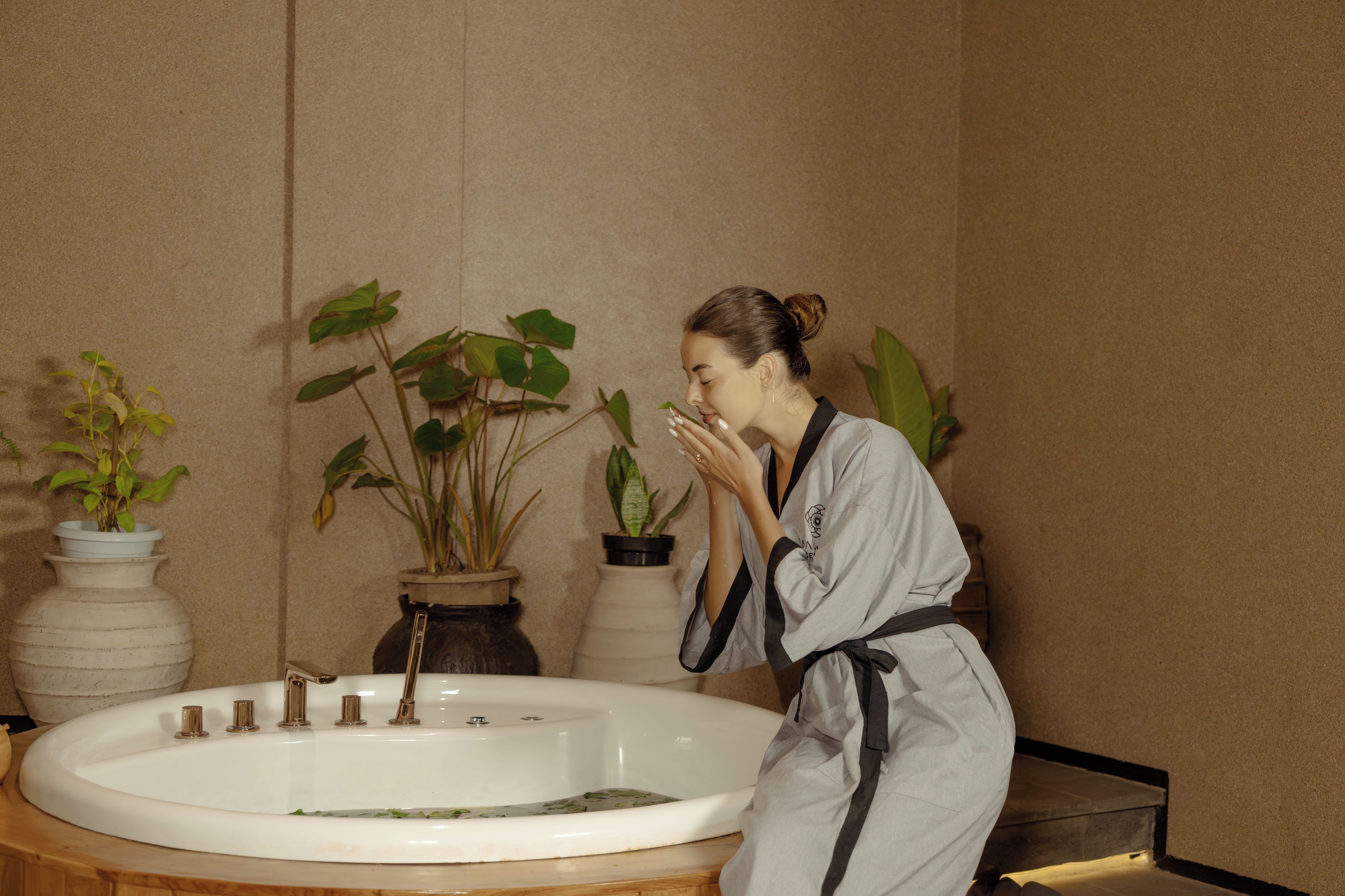 A person in a robe by a bathtub with multiple faucets, surrounded by potted plants.