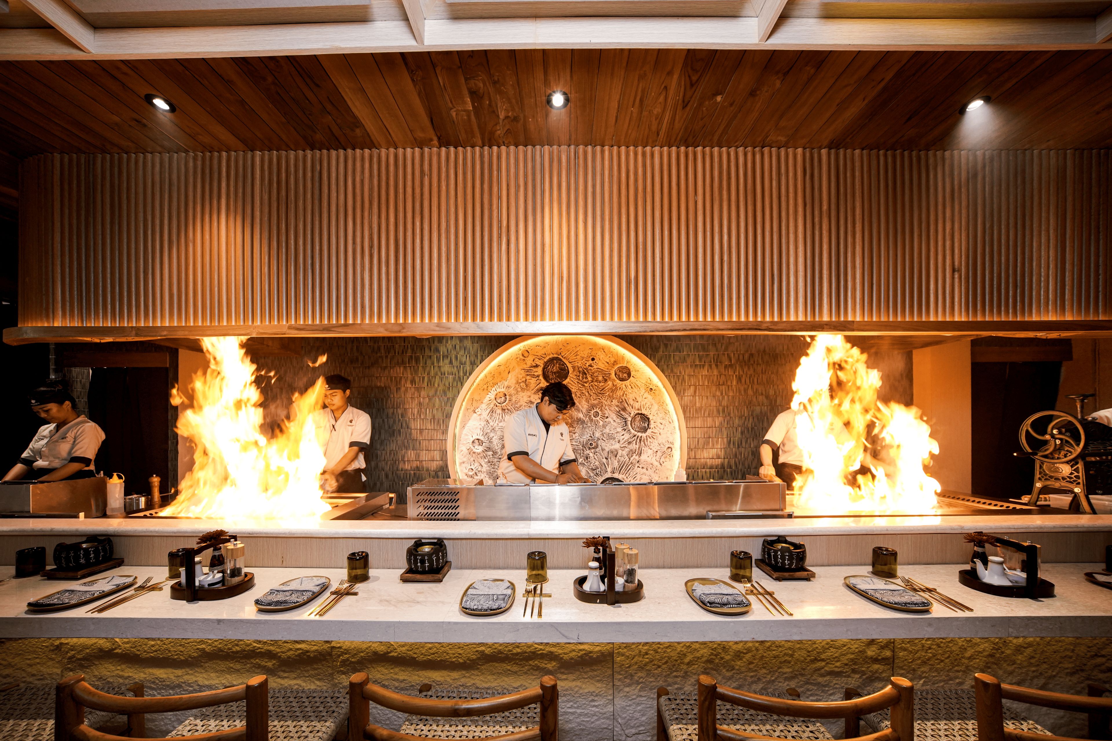 Un restaurante con una barra alargada, techo de madera y cocineros realizando una demostración de cocina con fuego.