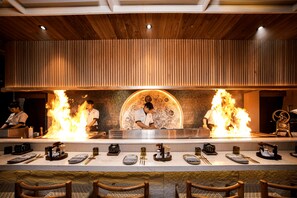 A restaurant with a long counter, wooden ceiling, and chefs performing a fire-based cooking demonstration. - Sini Vie Resort & Spa Seminyak by Ini Vie Hospitality (Keroboken Kelod)