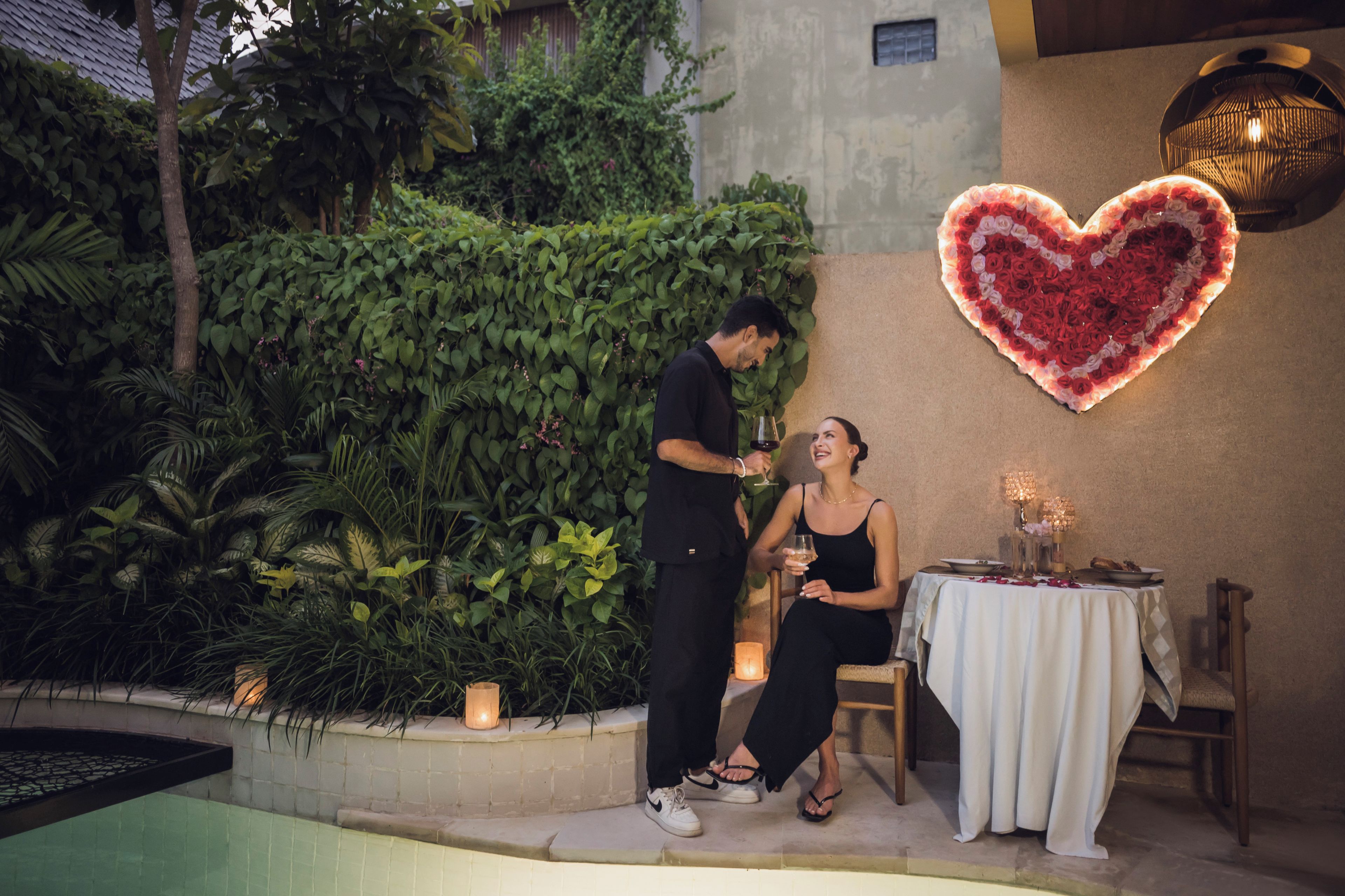 A couple standing by a table with a heart-shaped light fixture above, surrounded by greenery and a pool.