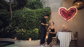 A couple standing by a table with a heart-shaped light fixture above, surrounded by greenery and a pool.