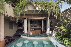 Un área de piscina con asientos de piedra, un bar con estantes llenos de botellas y una escalera que lleva a un nivel superior cubierto.