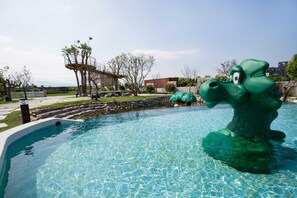 Ein Swimmingpool mit einer grünen Dinosaurier-Statue, einer überdachten Brücke und einem Spielplatz im Hintergrund.