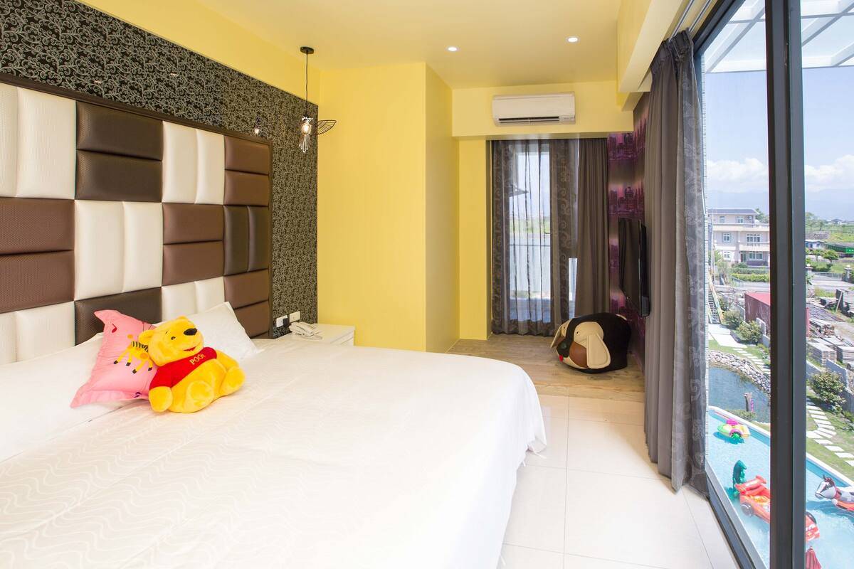 Una habitación de hotel con una cama grande, un peluche y vista a una piscina.