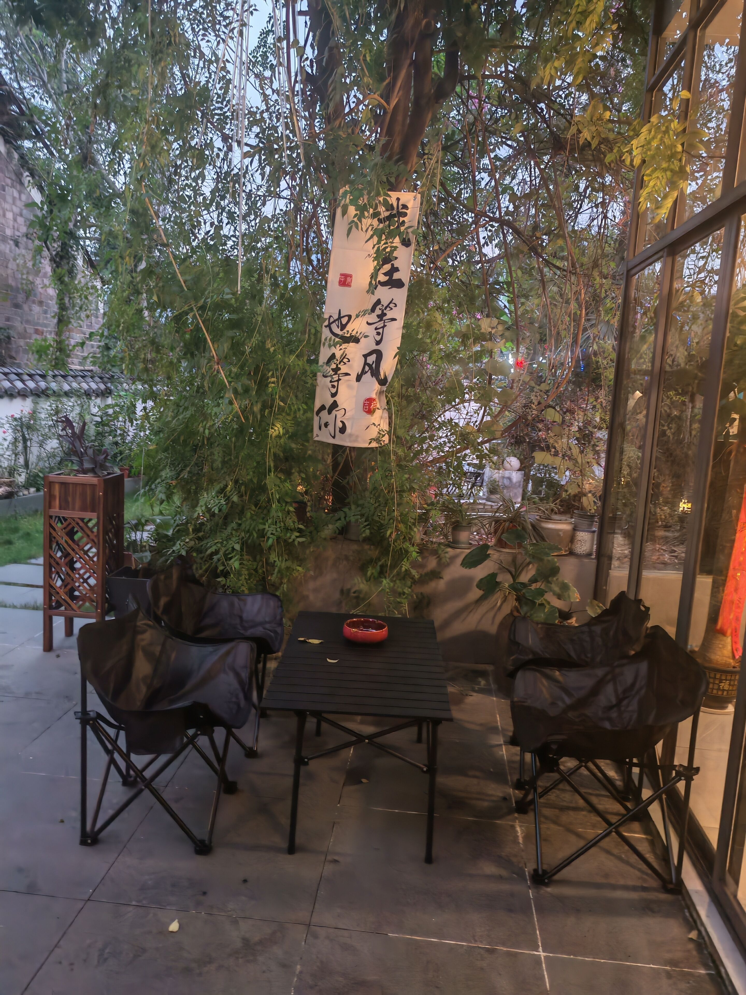 An outdoor seating area with a table and chairs, surrounded by greenery and a hanging banner with Chinese characters.