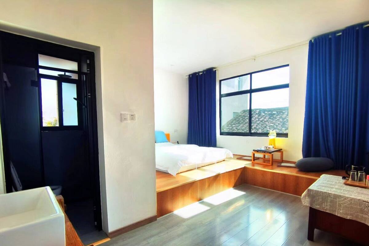 Une chambre d’hôtel avec un lit, une petite table avec une lampe, une fenêtre avec des rideaux et un banc en bois.