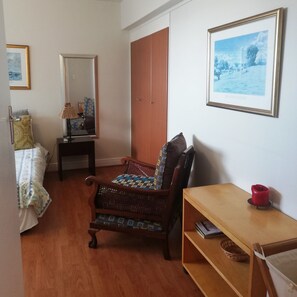 A room with a wooden floor, a bed with patterned bedding, a chair with a cushion, a wooden table with a red object, and a framed picture on the wall. - Villa Marina (Cape Town)