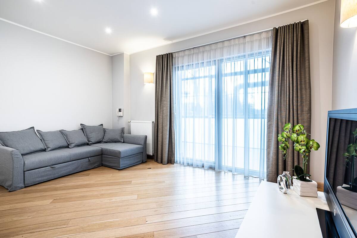 Una sala moderna con un sofá seccional gris, piso de madera, un televisor de pantalla plana y un ventanal con cortinas.