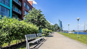 Een pad bekleed met banken en bomen, dat leidt naar een waterpartij met een brug op de achtergrond.
