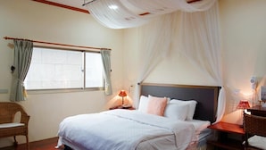 Une chambre à coucher avec un grand lit, deux tables de chevet avec des lampes, un fauteuil en osier, une fenêtre avec des rideaux et un baldaquin au-dessus du lit.
