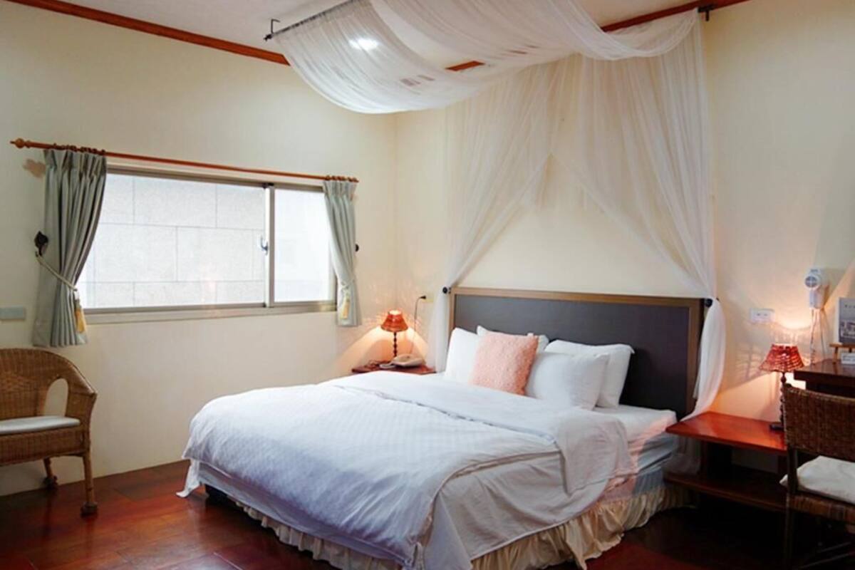 A bedroom with a large bed, two bedside tables with lamps, a wicker chair, a window with curtains, and a canopy over the bed.