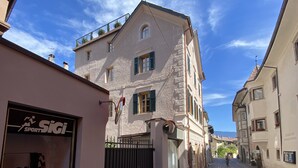 A cobblestone street lined with traditional buildings, a storefront with a sign reading "Sigi Sport," and a clear blue sky.