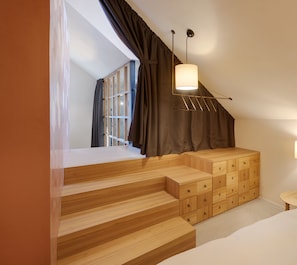 Un dormitorio con una escalera de madera, una cama con ropa blanca, una mesita de noche con lámpara y una ventana con cortinas.