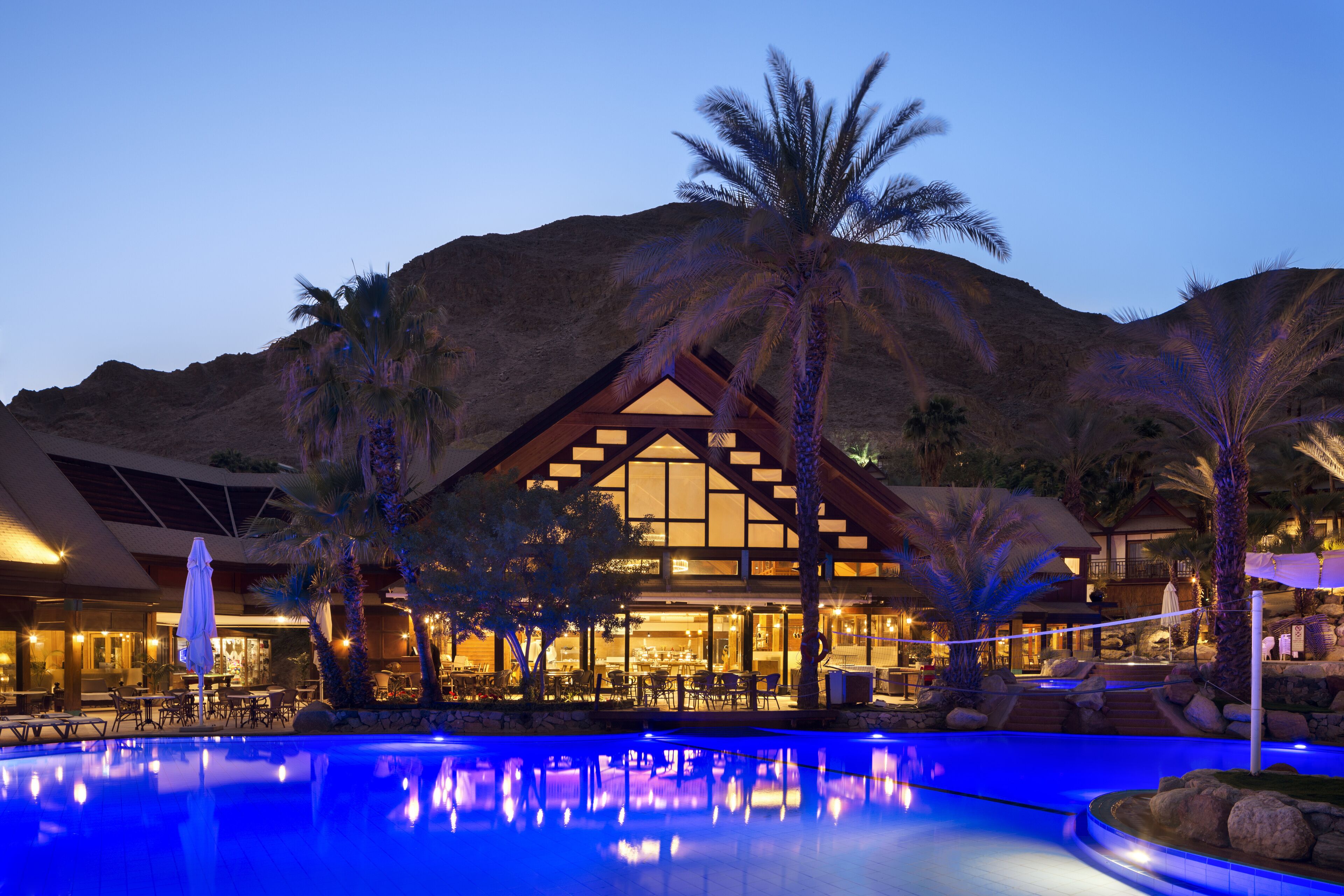 A resort with a swimming pool, outdoor dining area, and a large building with a distinctive roof structure.