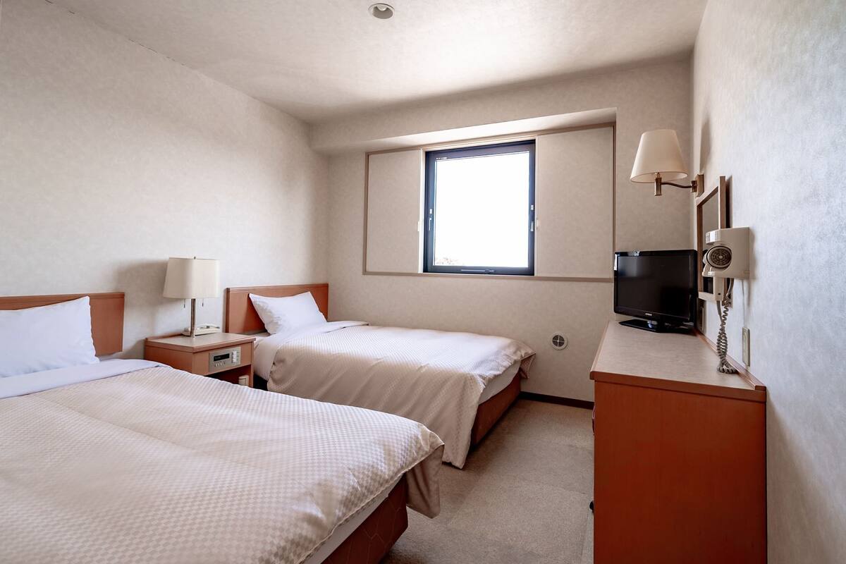 A hotel room with two beds, a television, a desk with a lamp, and a window.