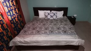 A neatly made bed with a floral bedspread and two pillows, a bedside table with a remote control, and patterned curtains on the walls. - Peemens Guesthouse (Ngleshi Amanfro)