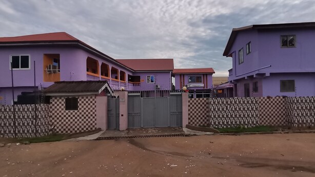 A gated residential area with purple houses and a checkered fence. - Peemens Guesthouse (Ngleshi Amanfro)