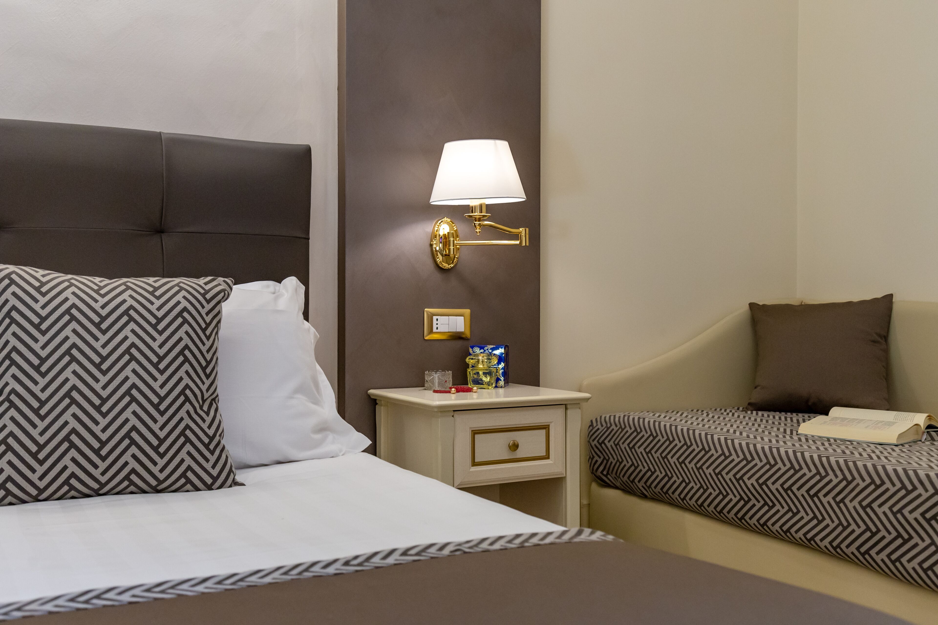 A hotel room with a large bed, two smaller beds, a bedside table with a lamp, and a nightstand with a book and a glass.