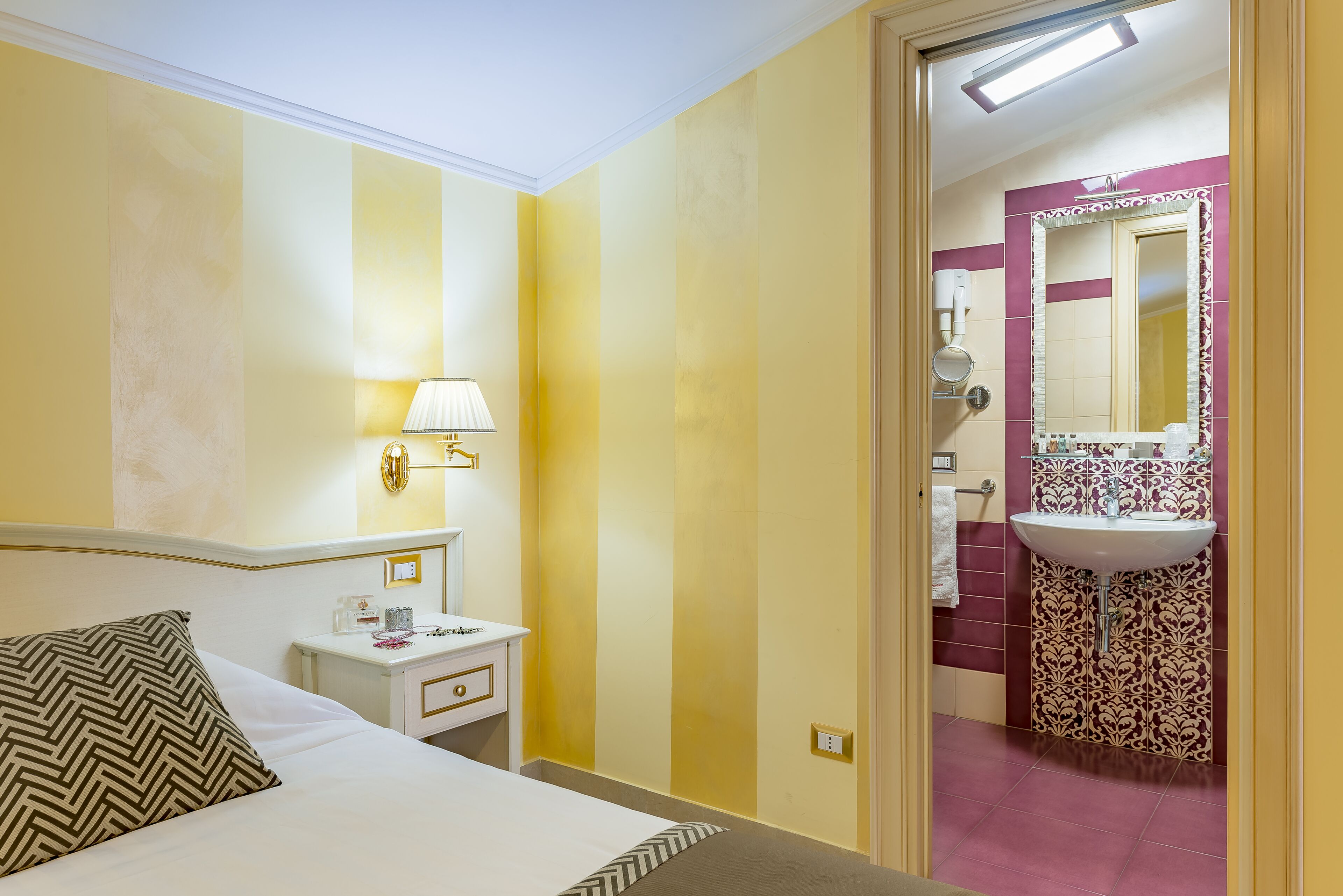Quarto de hotel com cama, mesa de cabeceira, abajur e banheiro com pia e espelho.