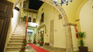 A grand staircase with a red carpet leading to a balcony, surrounded by potted plants and a fire extinguisher. - Vittorio Emanuele Boutique Hotel (Sciacca)