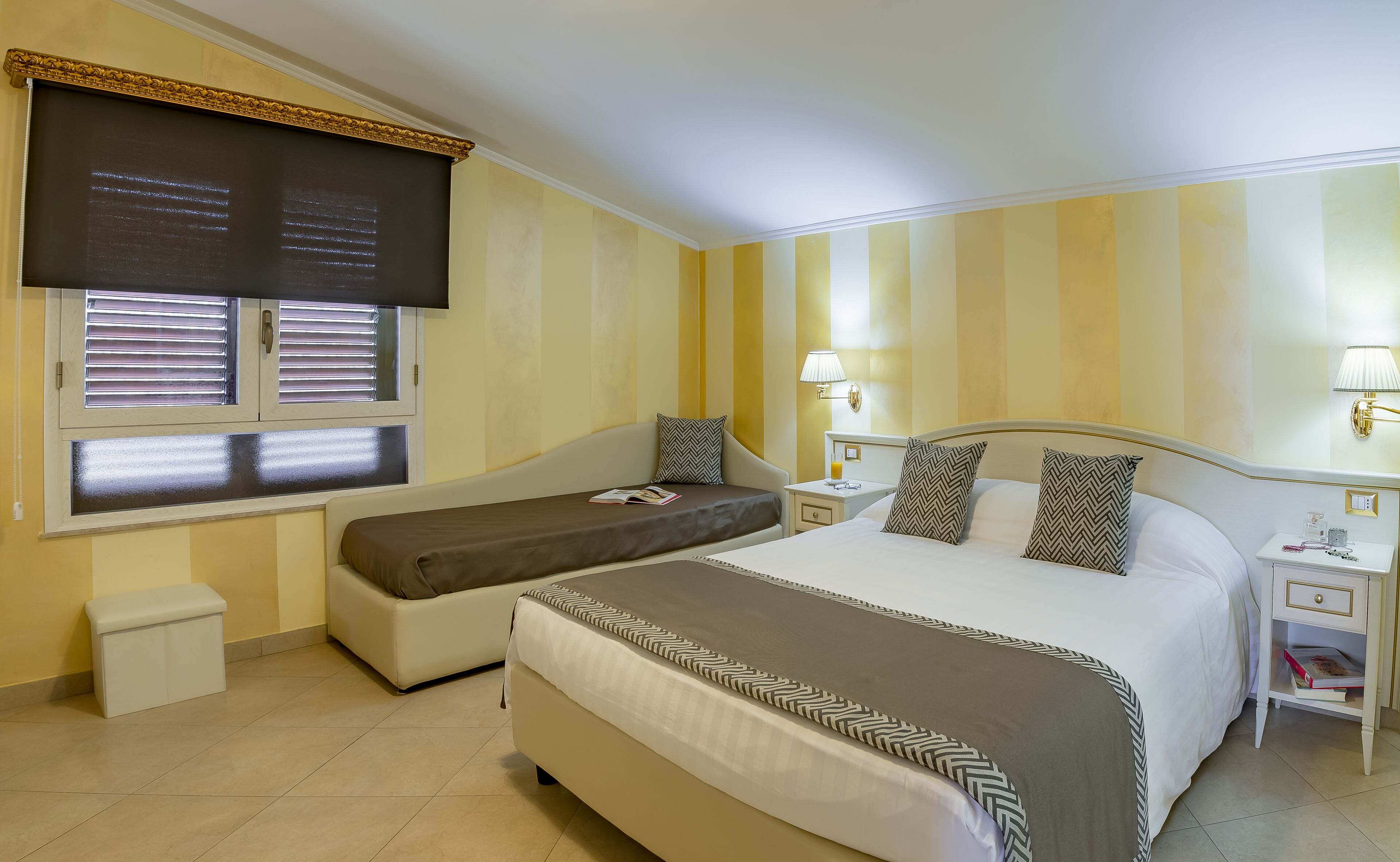 A hotel room with a large bed, a smaller sofa, a bedside table with a lamp, and a window with blinds.