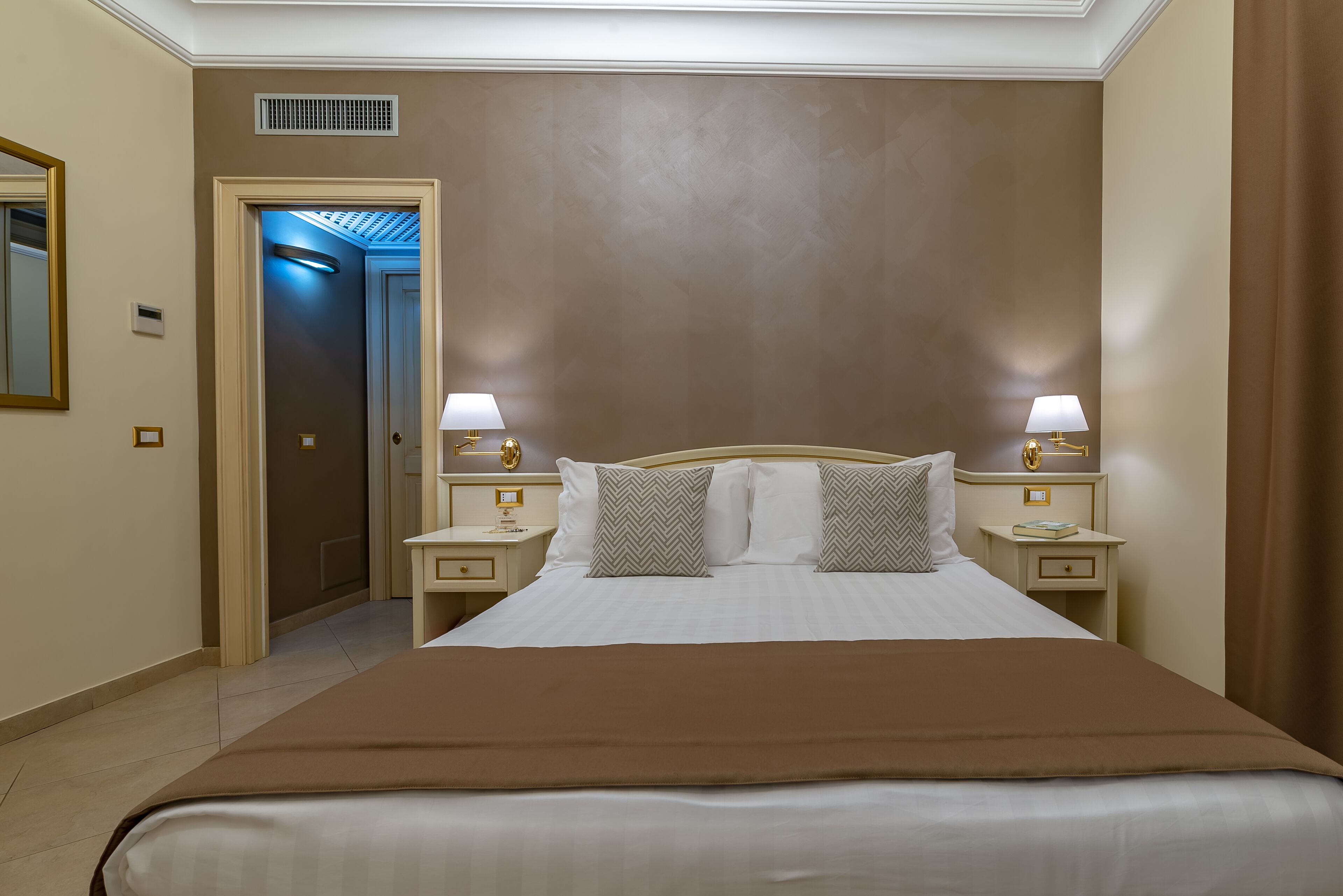 A hotel room with a large bed, two bedside tables with lamps, a mirror, and a door leading to another room.