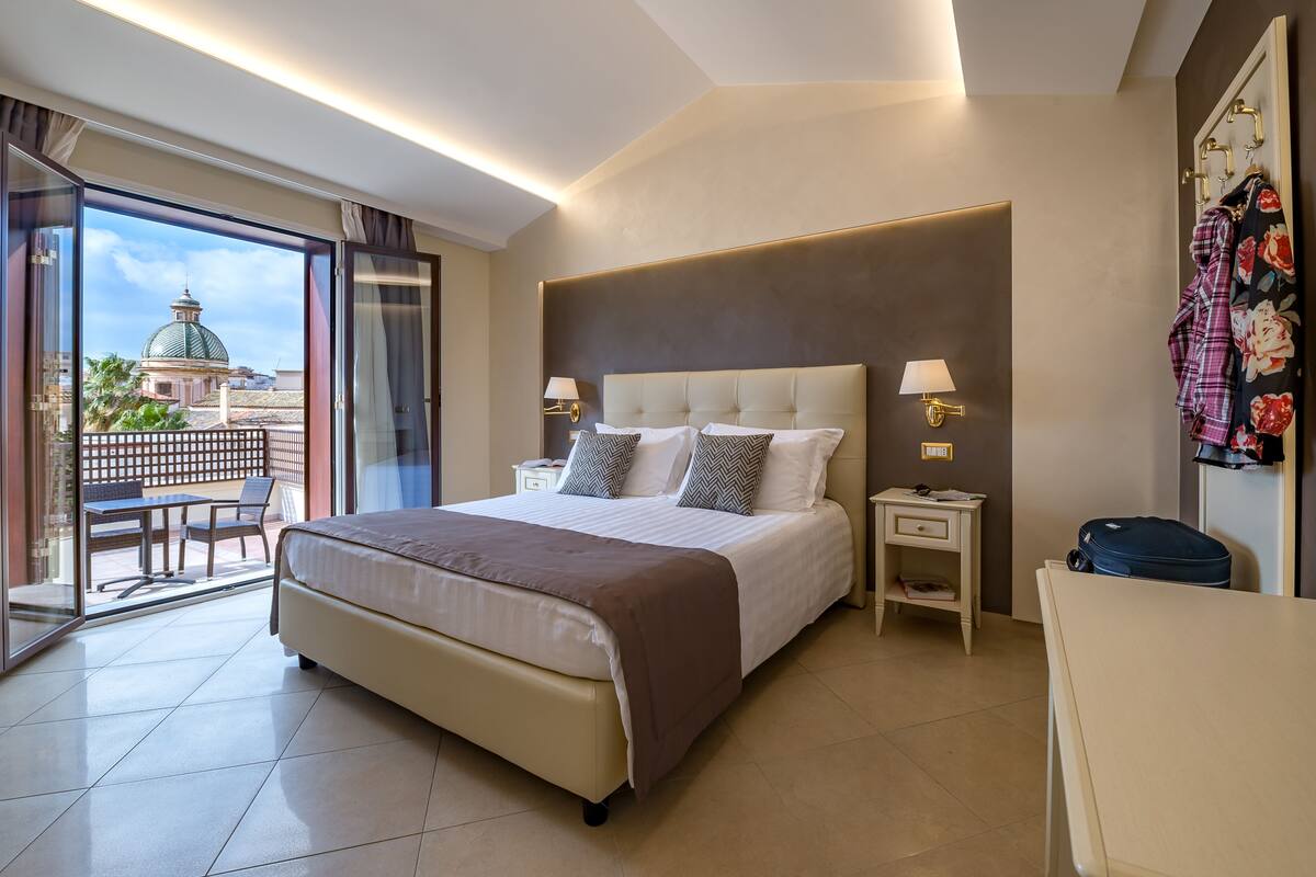 A hotel room with a large bed, two bedside tables, a desk, and a chair. There is a balcony with a view of a building and palm trees.